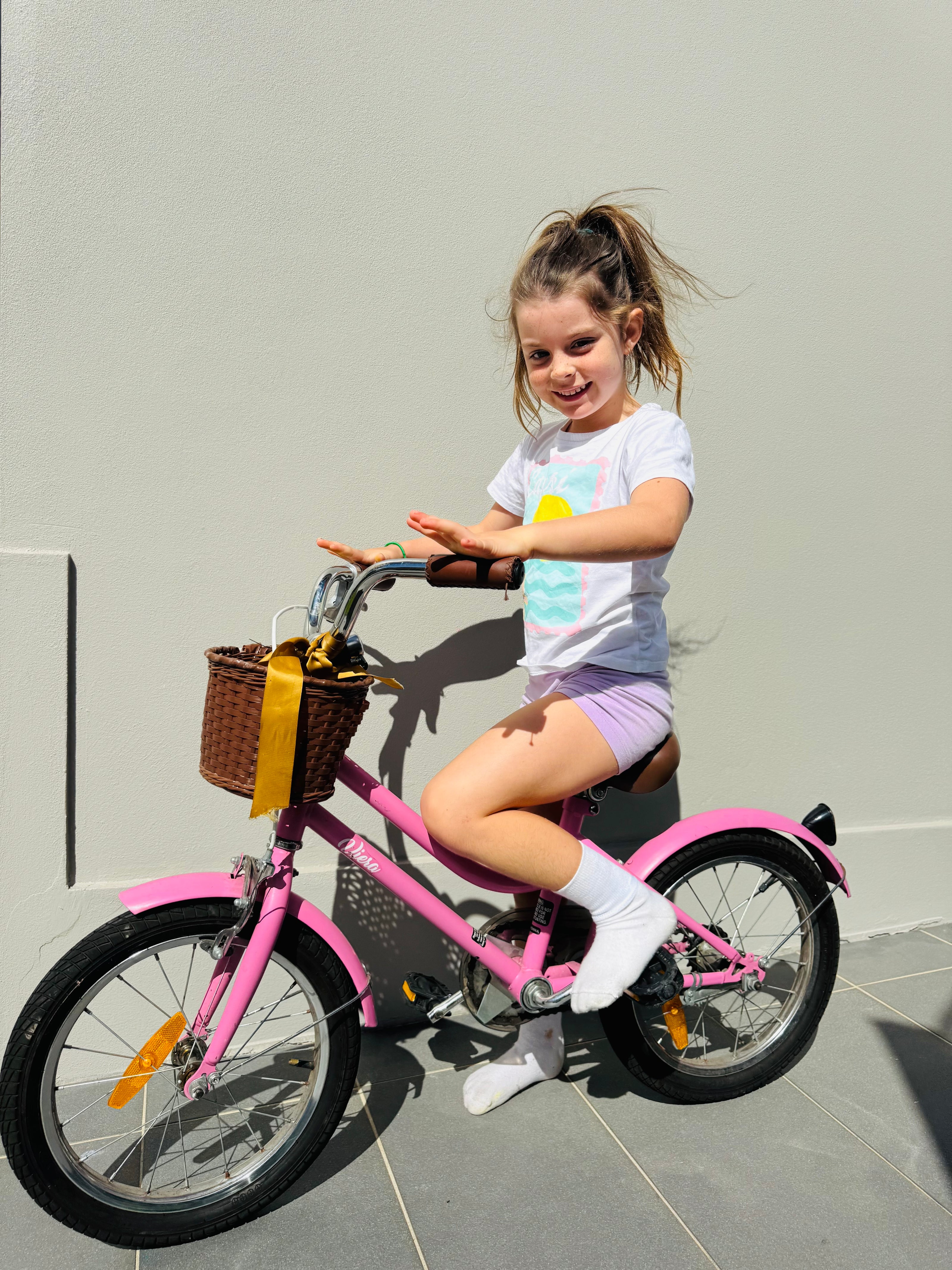 Young girl sitting on a pink children's bicycle against a plain wall.
